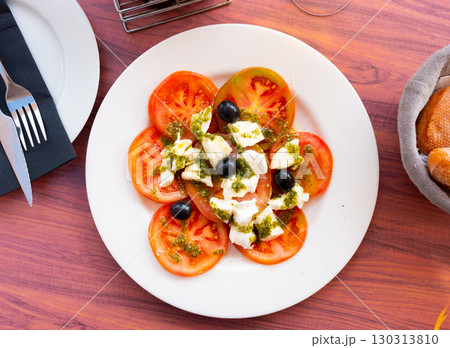 Appetizing salad of fresh ripe tomatoes with green pesto sauce and decorated with black olives. Italian cuisine Appetizing salad of fresh ripe tomatoes with green pesto sauce and decorated with black olives. Italian cuisine 130313810