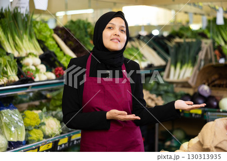 Female seller in a hijab and an apron offers to buy vegetables and fruits in the grocery department of supermarket 130313935