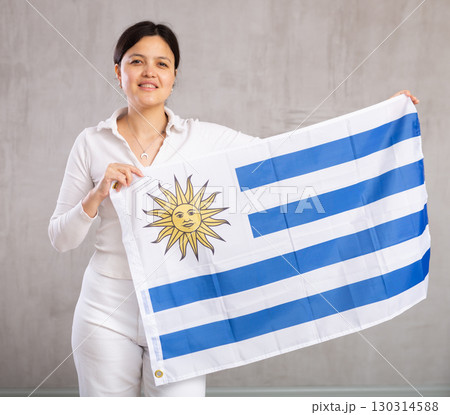 Reliant positive young woman holds big national flag of Uruguay with confident smile showing teeth Reliant positive young woman holds big national flag of Uruguay with confident smile showing teeth 130314588