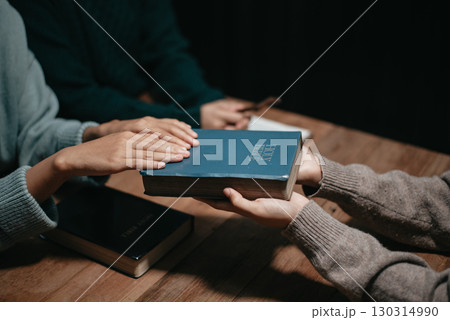 Group of Christians sit together and pray around a wooden table with blurred open Bible pages in their homeroom. Group of Christians sit together and pray around a wooden table with blurred open Bible pages in their homeroom. 130314990