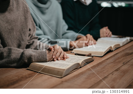 Group of Christians sit together and pray around a wooden table with blurred open Bible pages in their homeroom. Group of Christians sit together and pray around a wooden table with blurred open Bible pages in their homeroom. 130314991