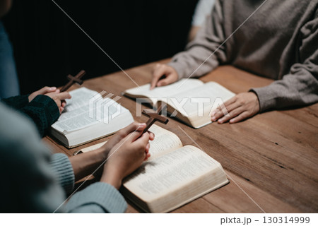 Group of Christians sit together and pray around a wooden table with blurred open Bible pages in their homeroom. 130314999