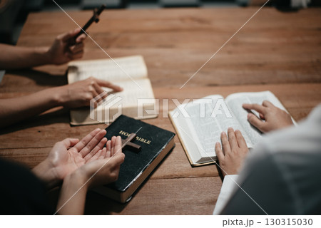 Group of Christians sit together and pray around a wooden table with blurred open Bible pages in their homeroom. 130315030