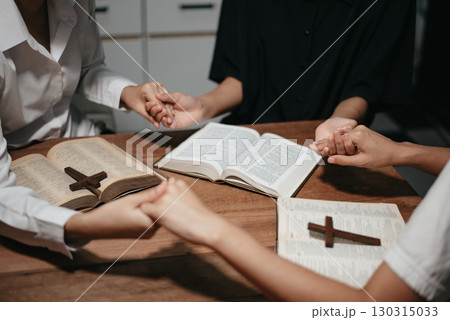Group of Christians sit together and pray around a wooden table with blurred open Bible pages in their homeroom. Group of Christians sit together and pray around a wooden table with blurred open Bible pages in their homeroom. 130315033