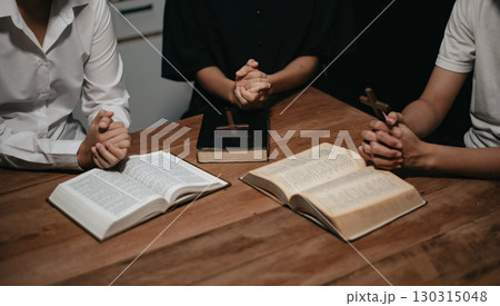 Group of Christians sit together and pray around a wooden table with blurred open Bible pages in their homeroom. Group of Christians sit together and pray around a wooden table with blurred open Bible pages in their homeroom. 130315048