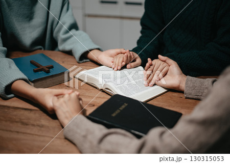 Group of Christians sit together and pray around a wooden table with blurred open Bible pages in their homeroom. 130315053