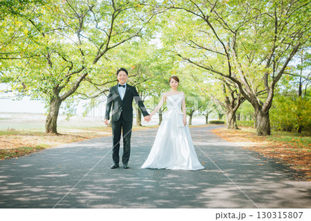 公園の並木道を手を繋いで歩くドレス姿のカップル（ウェディング・ウェディングフォト・新郎新婦） 130315807