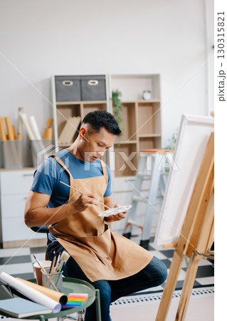 Asian male painter do artwork in art workshop, painting supplies, oil pastels, two canvas easel, creative space with paintbrush 130315821