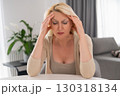 Middle aged blonde woman sitting at wooden table in cozy living room, leaning on elbows and pressing hands to temples, experiencing throbbing headache and fatigue 130318134