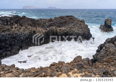 Waves crashing against rugged black volcanic rocks along the coast of La Graciosa, Lanzarote, with a view of the sea. 130319343