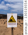 A warning sign showing the risk of falling is posted on a sandy beach with the ocean and blue sky in the background. 130319354