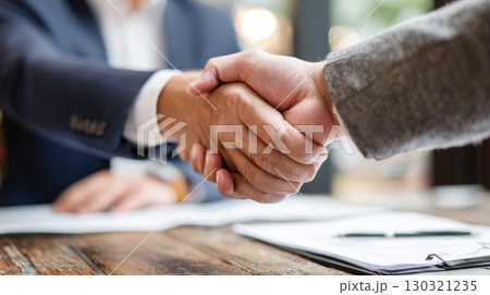Two business people shaking hands over wooden table with documents, expressing agreement and cooperation in professional setting Two business people shaking hands over wooden table with documents, expressing agreement and cooperation in professional setting 130321235