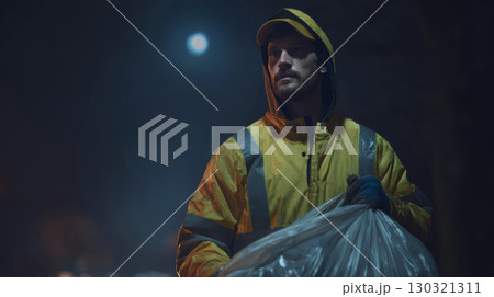 Man in yellow raincoat holding garbage bag at night with full moon in background, showing determination and focus 130321311
