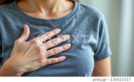 Woman holding hand on chest showing discomfort and pain, wearing casual gray shirt in soft natural light indoors Woman holding hand on chest showing discomfort and pain, wearing casual gray shirt in soft natural light indoors 130321517