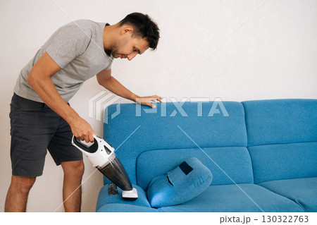 Cleaning service employee male using handheld vacuum cleaner to remove dust and dirt from blue sofa, emphasizing importance of cleanliness and hygiene in home maintenance. Cleaning service employee male using handheld vacuum cleaner to remove dust and dirt from blue sofa, emphasizing importance of cleanliness and hygiene in home maintenance. 130322763