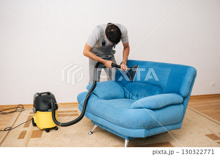 Wide shot of professional cleaning service worker removing dirt and stains from blue upholstered sofa using extraction washing vacuum cleaner in living room. Concept of domestic work, housekeeping. 130322771