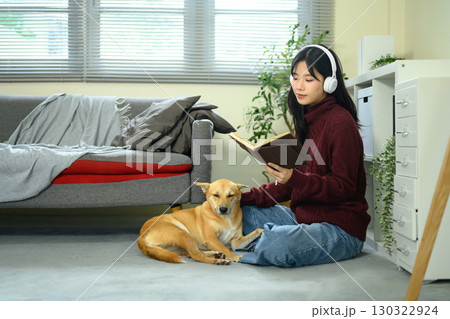 Young woman wearing headphones reading a book while sitting on the floor with her dog Young woman wearing headphones reading a book while sitting on the floor with her dog 130322924