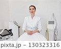 Portrait of confident female cosmetologist in white uniform sitting in aesthetic clinic. Professional skincare expert with clean background, beauty equipment, laser devices. Health and wellness 130323316