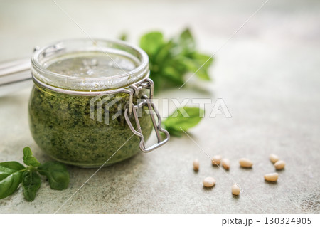 Fresh homemade basil pesto in glass jar with scattered pine nuts and basil leaves on speckled background. Food photography with soft focus and copy space 130324905