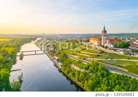 Aerial view captures Melnik City and its historic chateau perched above the serene Labe River. The landscape features lush greenery and tranquil waters during golden hour. Aerial view captures Melnik City and its historic chateau perched above the serene Labe River. The landscape features lush greenery and tranquil waters during golden hour. 130326081