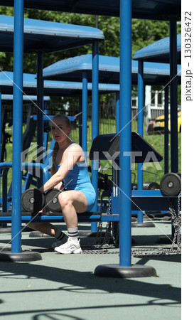 A woman in a blue athletic outfit is seated at an outdoor gym, lifting dumbbells over her head. This image highlights the dedication to strength training and the benefits of exercising in open-air 130326472