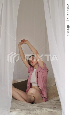 A woman stretches gracefully on a comfortable bed, basking in the calm of her surroundings. The photo reflects the beauty of slow mornings and prioritizing well-being in daily life. 130327502