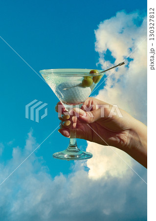 Hand holding martini glass with olives and golf ball against blue sky stylish surreal mood 130327812