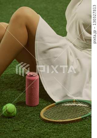 Woman in white dress sitting on grass with pink soda can and vintage tennis racket playful vibe 130327813