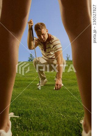 Male golfer crouching to place ball on grass seen through female legs focused confident mood Male golfer crouching to place ball on grass seen through female legs focused confident mood 130327827