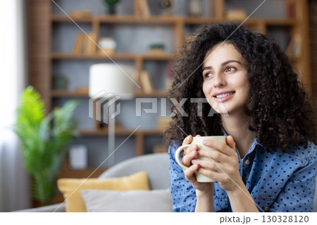 Young woman with curly hair holding a mug and relaxing on the sofa, enjoying a moment of peace and tranquility in her modern living room, smiling and looking away. Young woman with curly hair holding a mug and relaxing on the sofa, enjoying a moment of peace and tranquility in her modern living room, smiling and looking away. 130328120