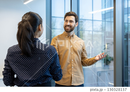 Businessman explaining work strategy to female colleague, gesturing with hands and smiling, during a meeting in a modern office space with large windows 130328137