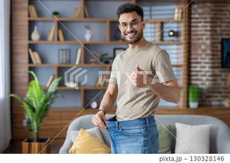 Happy man demonstrates impressive weight loss by showing his loose jeans while giving thumbs up. Person expresses joy and achievement in modern living room space, highlighting personal transformation Happy man demonstrates impressive weight loss by showing his loose jeans while giving thumbs up. Person expresses joy and achievement in modern living room space, highlighting personal transformation 130328146