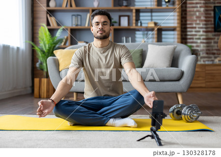 Indian young man doing online yoga at home, sitting on mat in lotus position with closed eyes in front of mobile phone. 130328178