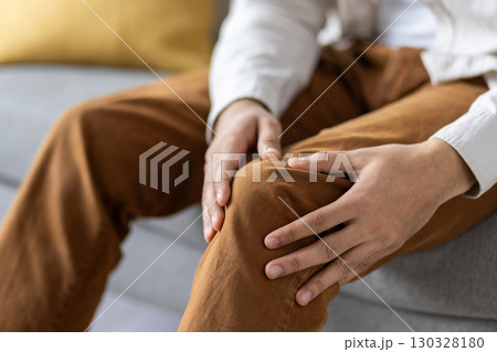 Person seated on sofa holding knee, indicating discomfort or pain. Captures concept of injury, health concern, or joint problem. Emphasizes human touch and care related to knee pain. Person seated on sofa holding knee, indicating discomfort or pain. Captures concept of injury, health concern, or joint problem. Emphasizes human touch and care related to knee pain. 130328180
