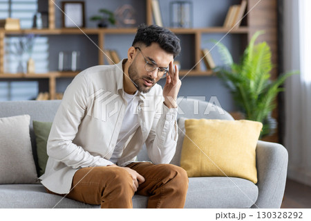 Young man with glasses experiencing headache sitting on sofa, displaying stress and fatigue. Casual setting at home, holding head in discomfort, wearing light shirt and glasses. 130328292