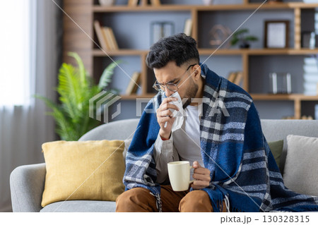 Young man sitting on sofa covered with blanket, holding hot drink and tissue. He appears sick, blowing nose, wrapped in warm blanket, trying to feel better at home during illness or cold season. Young man sitting on sofa covered with blanket, holding hot drink and tissue. He appears sick, blowing nose, wrapped in warm blanket, trying to feel better at home during illness or cold season. 130328315