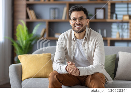 Smiling man with glasses relaxing on sofa in modern living room. Casual style with beige shirt and white t-shirt. Bright, cozy atmosphere reflects warmth and tranquility. Smiling man with glasses relaxing on sofa in modern living room. Casual style with beige shirt and white t-shirt. Bright, cozy atmosphere reflects warmth and tranquility. 130328344