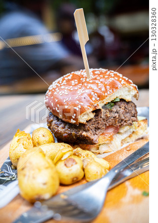 Juicy hamburger with sesame seed bun, garnished with fresh vegetables, served alongside crispy roasted potatoes on wooden platter 130328360