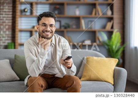 Smiling young man with glasses sitting comfortably on sofa holding TV remote control. Relaxed atmosphere, enjoying leisure and entertainment. Casual outfit with cozy home setting and warm lighting Smiling young man with glasses sitting comfortably on sofa holding TV remote control. Relaxed atmosphere, enjoying leisure and entertainment. Casual outfit with cozy home setting and warm lighting 130328440
