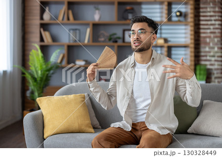Young man sitting on couch using hand fan to cool down in warm home environment. Expressive gestures indicate relaxation and casual setting during hot day. 130328449