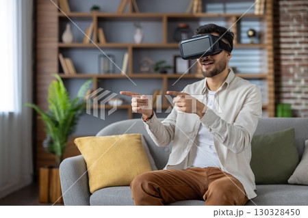 Young man immersed in virtual reality using VR headset, sitting on sofa and interacting with virtual environment. Smiling enthusiastically, experiencing cutting-edge technology excitement 130328450