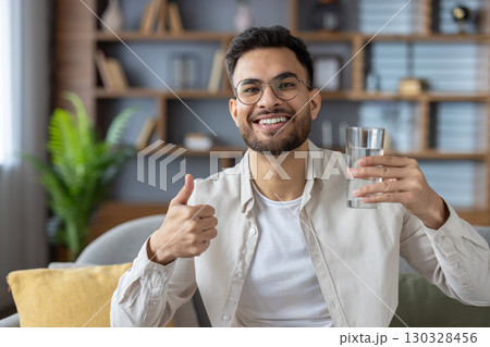 Smiling man with glasses holding glass of water showing thumbs-up. He sits comfortably in modern living room conveying positivity, health, and hydration. 130328456