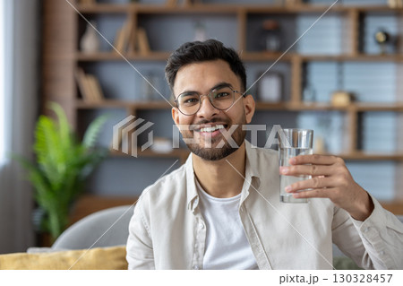 Smiling man wearing glasses holding glass of water, exuding confidence and wellness. Indoor scene suggests relaxed, healthy lifestyle. Ideal for concepts of hydration, positivity, and happiness. 130328457