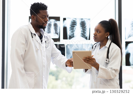 Male and female doctors discussing patient diagnosis in a clinical environment with x-rays visible. Professional medical team conferring about healthcare needs in modern clinic near window. Male and female doctors discussing patient diagnosis in a clinical environment with x-rays visible. Professional medical team conferring about healthcare needs in modern clinic near window. 130329252