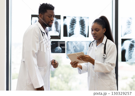 Male and female doctors discussing patient diagnosis, consulting x-ray images at clinic window. Collaborative healthcare professionals analyzing medical information to provide accurate treatment plan. Male and female doctors discussing patient diagnosis, consulting x-ray images at clinic window. Collaborative healthcare professionals analyzing medical information to provide accurate treatment plan. 130329257