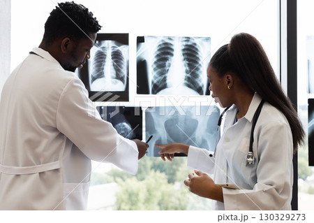 Male and female doctors analyzing x-rays together in a well-lit clinic environment near window. They are collaborating on reviewing medical images to determine diagnosis or treatment. 130329273