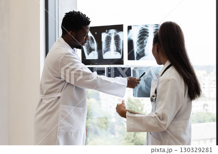 Male and female doctors of diverse backgrounds examining chest X-ray images by window. Collaborative medical consultation in modern clinic setting, emphasizing teamwork, analysis 130329281