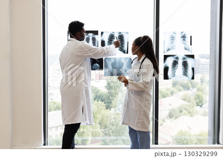 Medical professionals, male and female, reviewing x-ray diagnoses near a bright clinic window. Collaborative discussion over radiological findings indicates teamwork for accurate analysis. 130329299