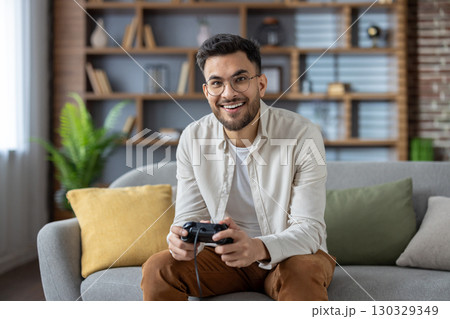 Hispanic man sitting on comfortable sofa holding gaming controller, engaging in leisure activity. Casual setting reflects enjoyment, relaxation, and entertainment at home. Hispanic man sitting on comfortable sofa holding gaming controller, engaging in leisure activity. Casual setting reflects enjoyment, relaxation, and entertainment at home. 130329349