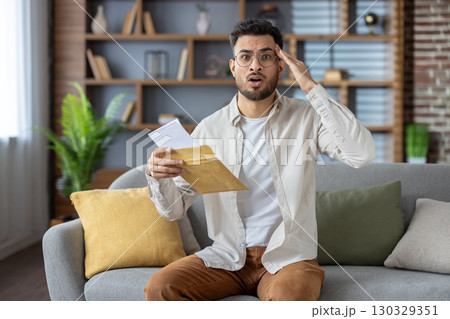 Portrait of shocked young Indian man sitting on sofa at home. holds the envelope with the received letter in his hand, looks worriedly into the camera. Portrait of shocked young Indian man sitting on sofa at home. holds the envelope with the received letter in his hand, looks worriedly into the camera. 130329351